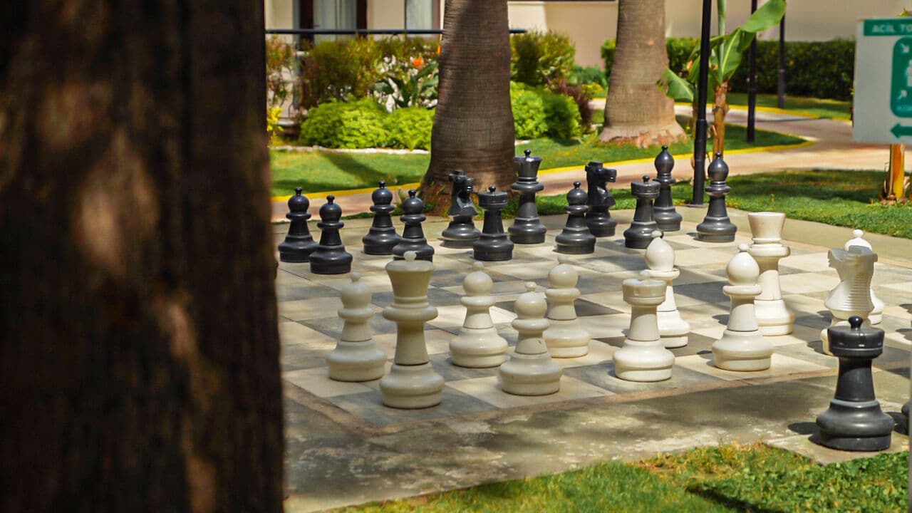 A large outdoor chessboard set up on a paved area with trees and greenery in the background.