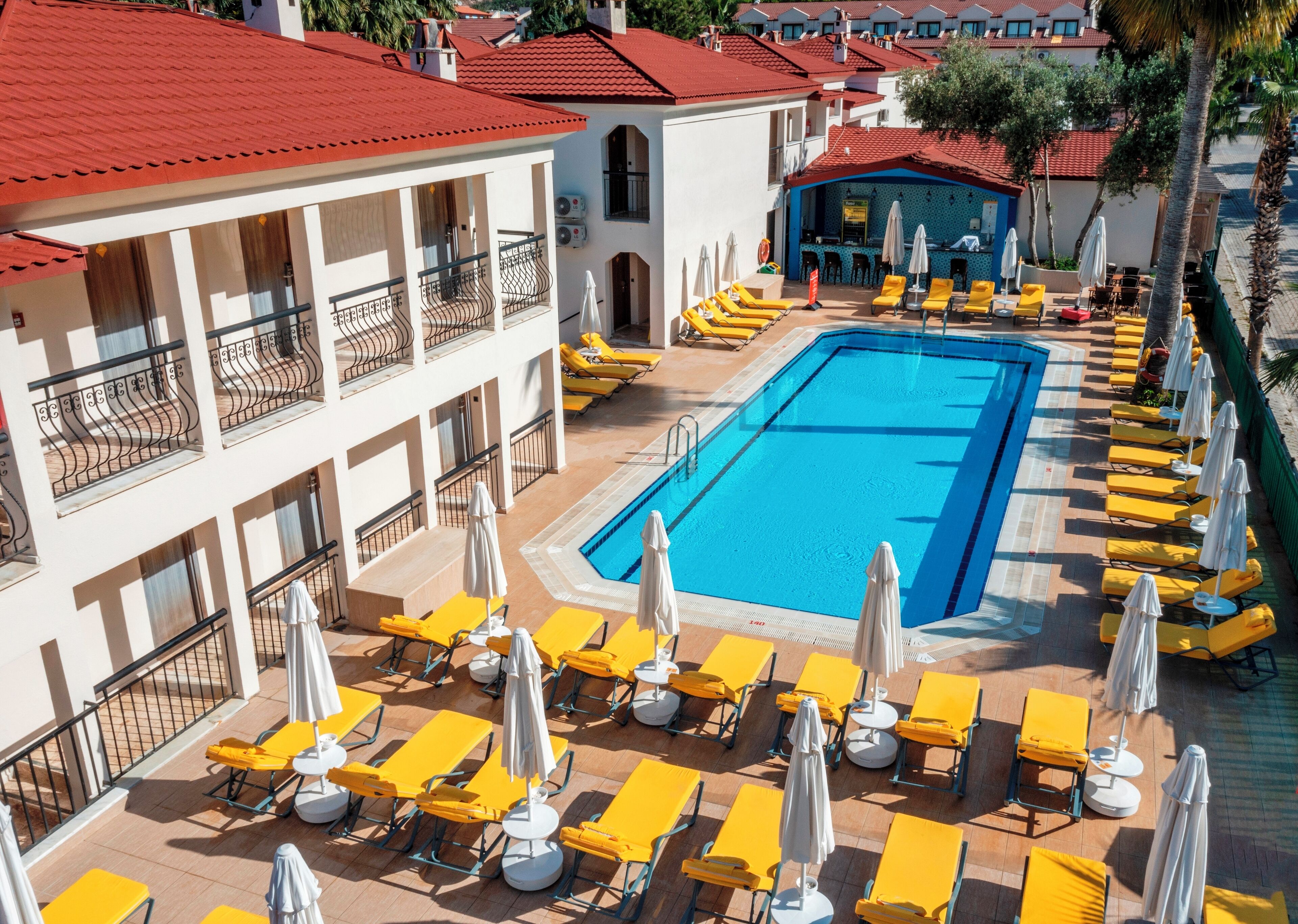 Ein Hotel mit Schwimmbecken, umgeben von gelben Liegestühlen und Sonnenschirmen, mit roten Ziegeldächern und weißen Wänden.