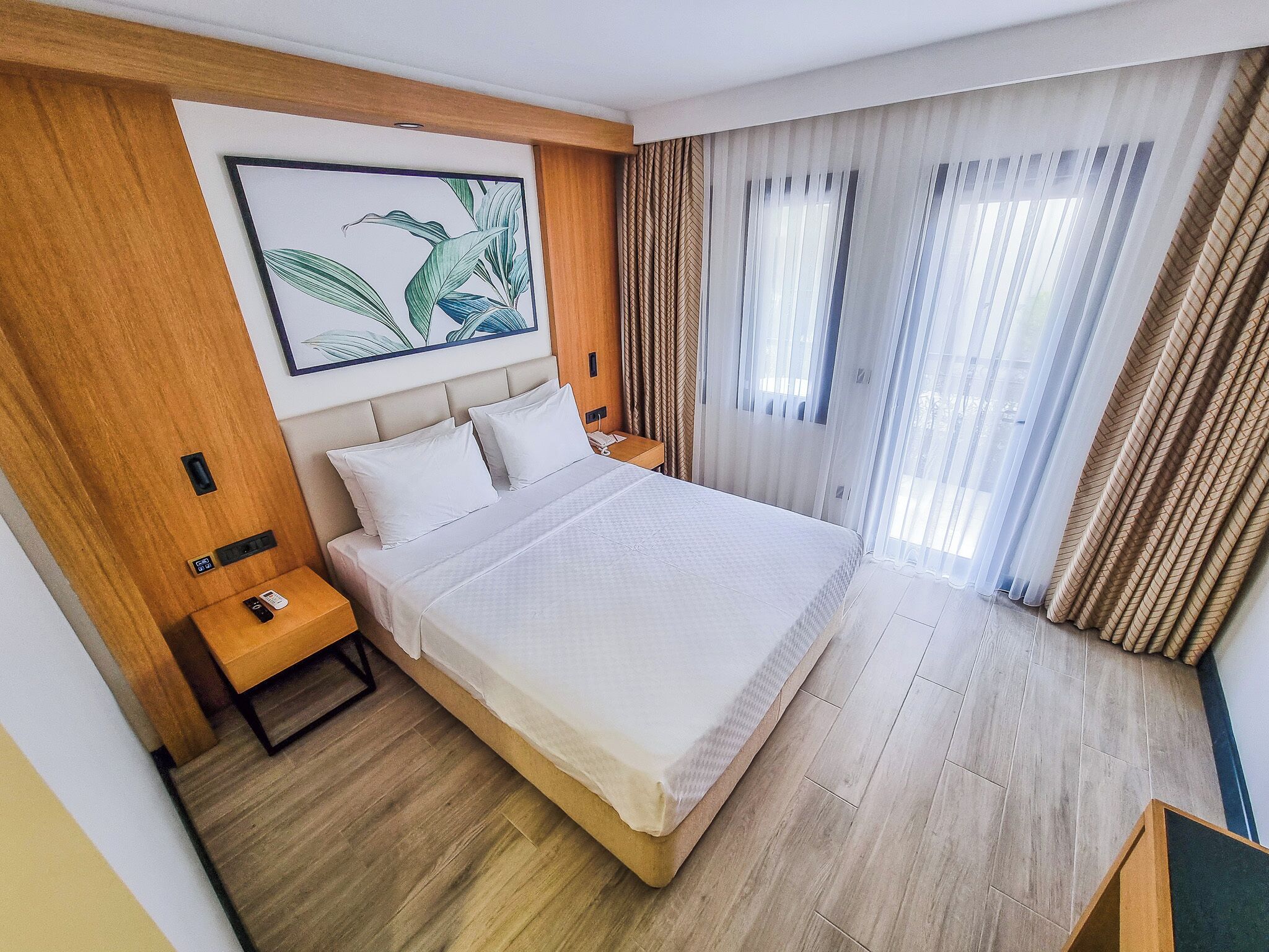 Habitación de hotel con una cama grande, suelo de madera, una mesita de noche y un teléfono montado en la pared, además de un ventanal con cortinas.