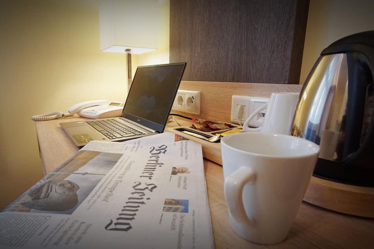 Ein Hotelzimmer mit einem Laptop, einer Zeitung, einer Kaffeetasse, einem Telefon und einem Wasserkocher auf dem Tisch.