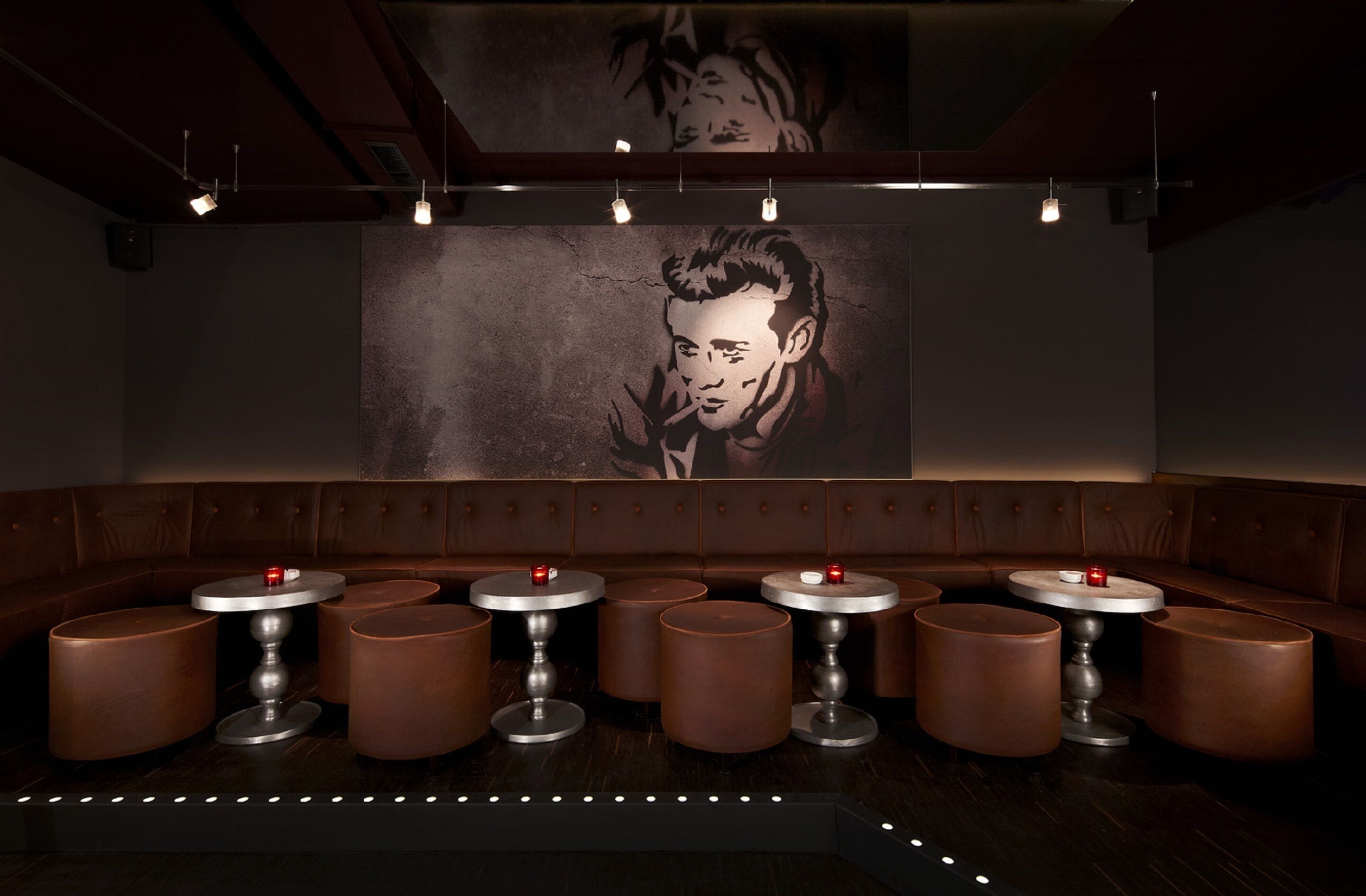 A dimly lit bar area with a large mural on the wall, multiple round tables with stools, and a row of cushioned seating.