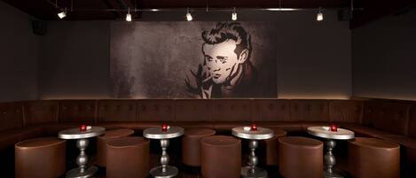 A dimly lit bar area with a large mural on the wall, multiple round tables with stools, and a row of cushioned seating.