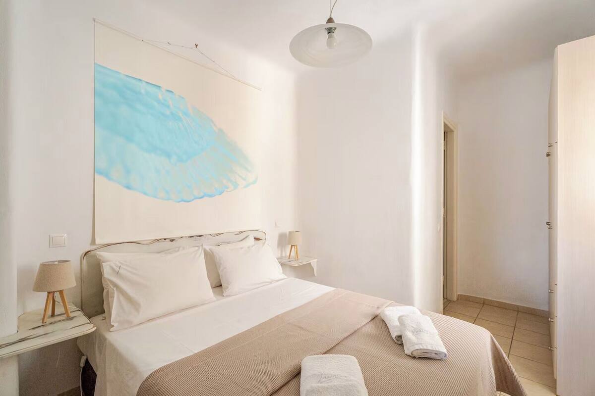 Une chambre à coucher avec un lit, deux tables de chevet avec des lampes, un luminaire suspendu et un grand tableau abstrait accroché au mur.