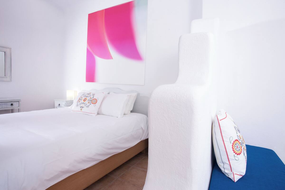 Un lit bien fait, avec du linge de lit blanc et des coussins décoratifs, une table de chevet avec une lampe, et un grand tableau abstrait accroché au mur.