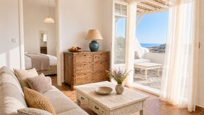A cozy living room with a sofa, a wooden cabinet, a table with a bowl and flowers, and a view of the sea through large windows. - Yakinthos Residence (Mykonos)