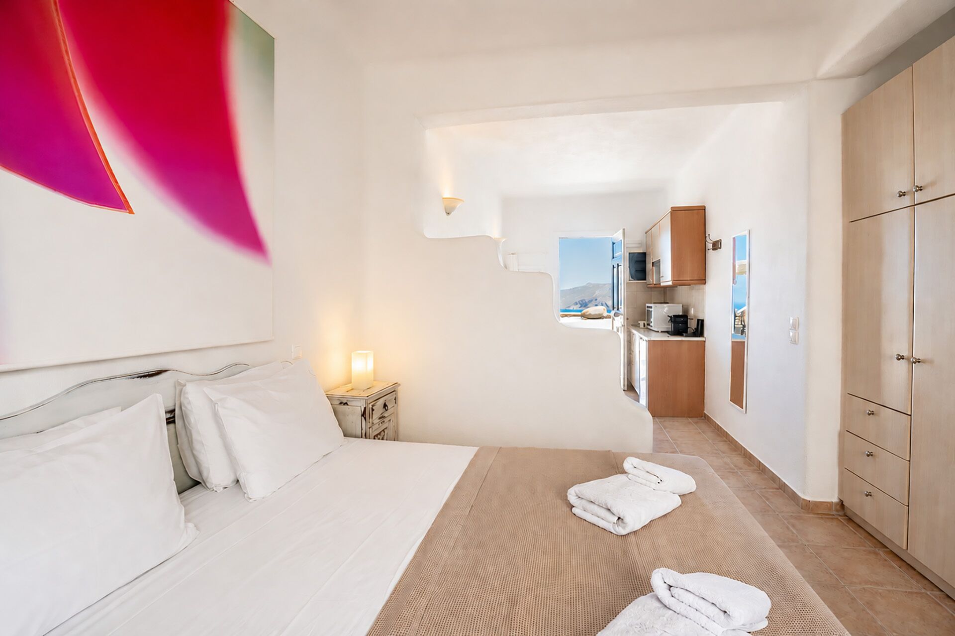 Habitación de hotel con cama, mesita de noche, vista a través de una puerta abierta y kitchenette.