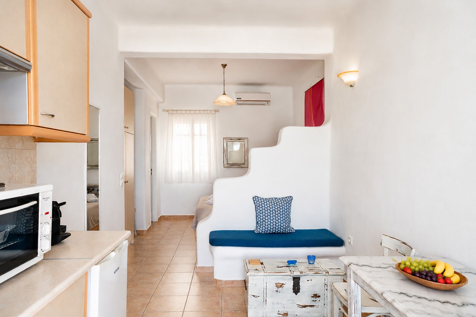 A compact kitchen with a microwave, a small refrigerator, and a dining area with a marble table and a bowl of fruit.