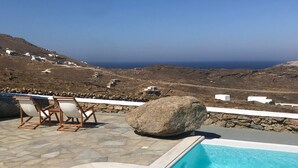 A pool with a stone deck, a large rock, and a view of a coastal landscape with buildings. - Yakinthos Residence (Mykonos)
