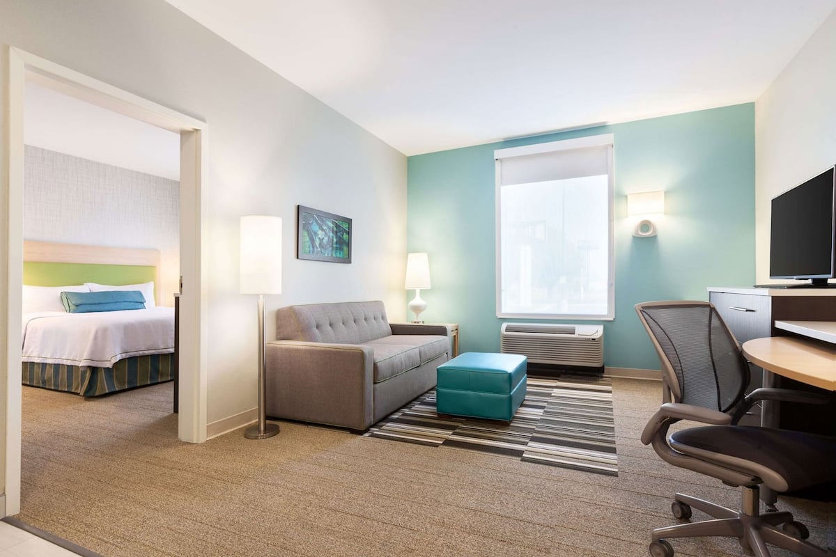 A hotel room with a bed, a sofa, a desk with a chair, a window with blinds, and a wall-mounted TV.