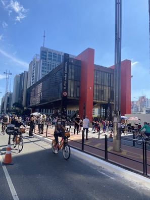 A busy urban street with cyclists and pedestrians, a prominent red building, and a clear blue sky. - Nomade In Arte e Hostel São Paulo (São Paulo)