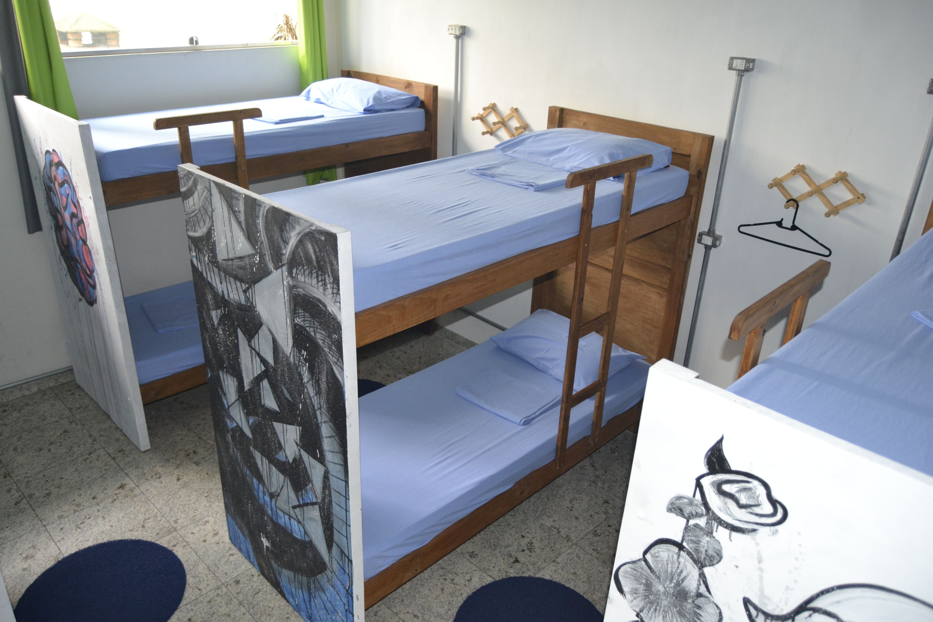 A dormitory room with bunk beds, a window with green curtains, a wall-mounted drying rack, and a poster on the wall.