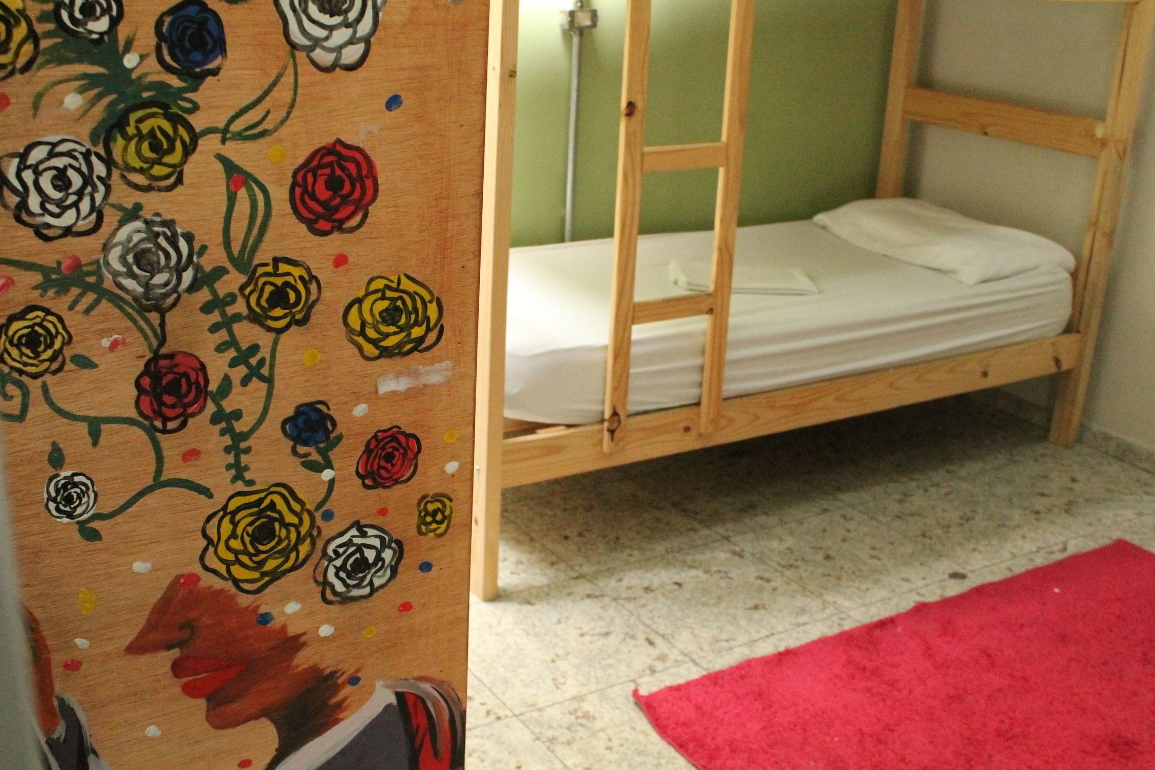 Una cama litera con estructura de madera y ropa de cama blanca, una alfombra roja y un cuadro floral en la pared.