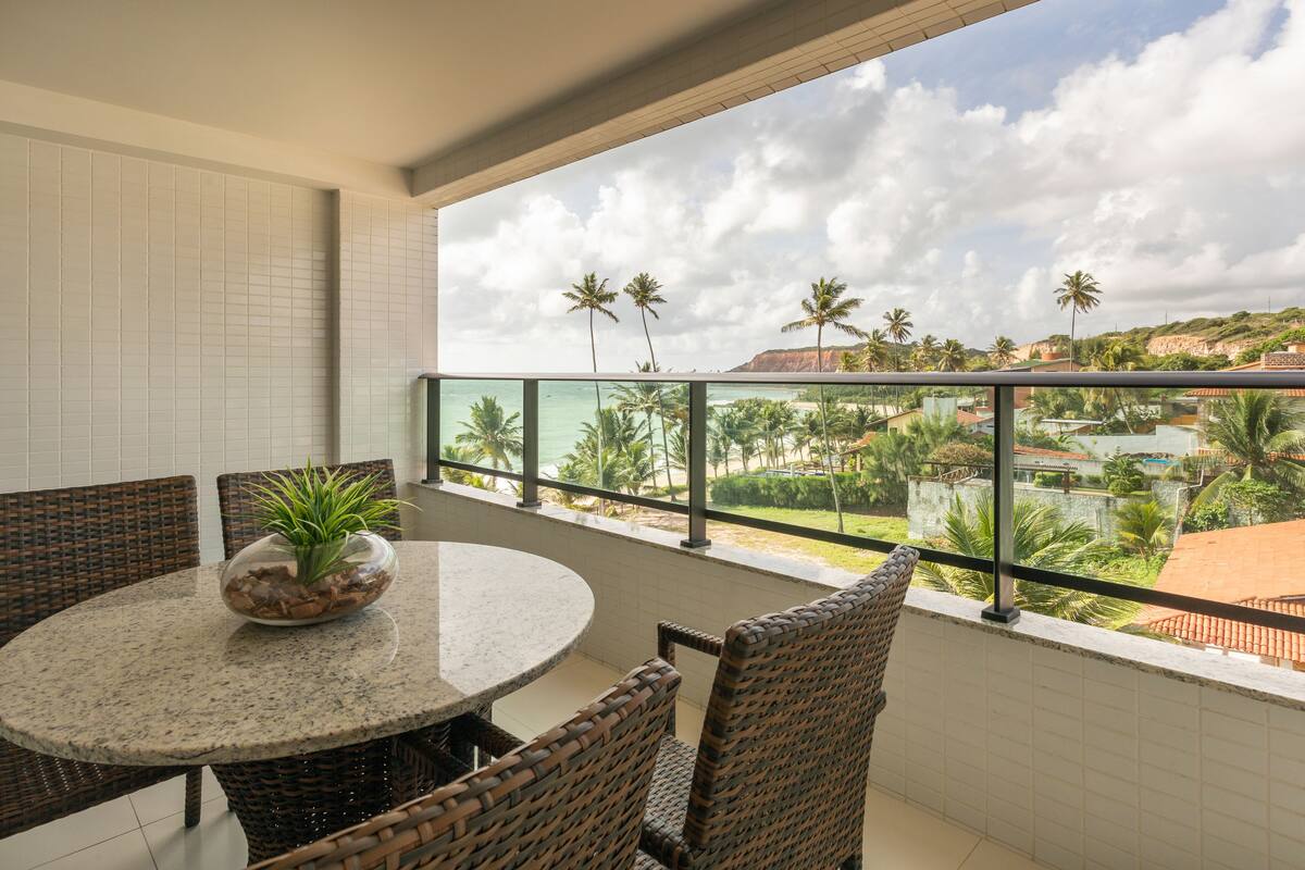 A balcony with a round table and wicker chairs, overlooking a resort area with palm trees and buildings.