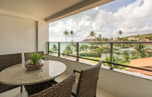 A balcony with a round table and wicker chairs, overlooking a resort area with palm trees and buildings. - Nord Life Tabatinga (João Pessoa)