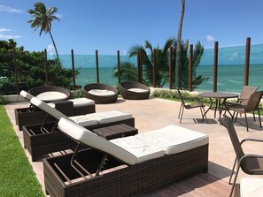 A patio area with wicker furniture, including lounge chairs and a table, overlooking a clear blue sea. - Nord Life Tabatinga (João Pessoa)