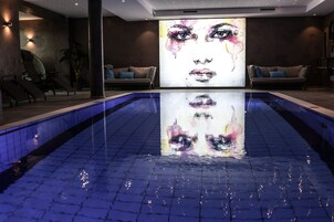 Una piscina interior moderna con una gran pantalla digital que muestra un rostro, rodeada de asientos y una planta.