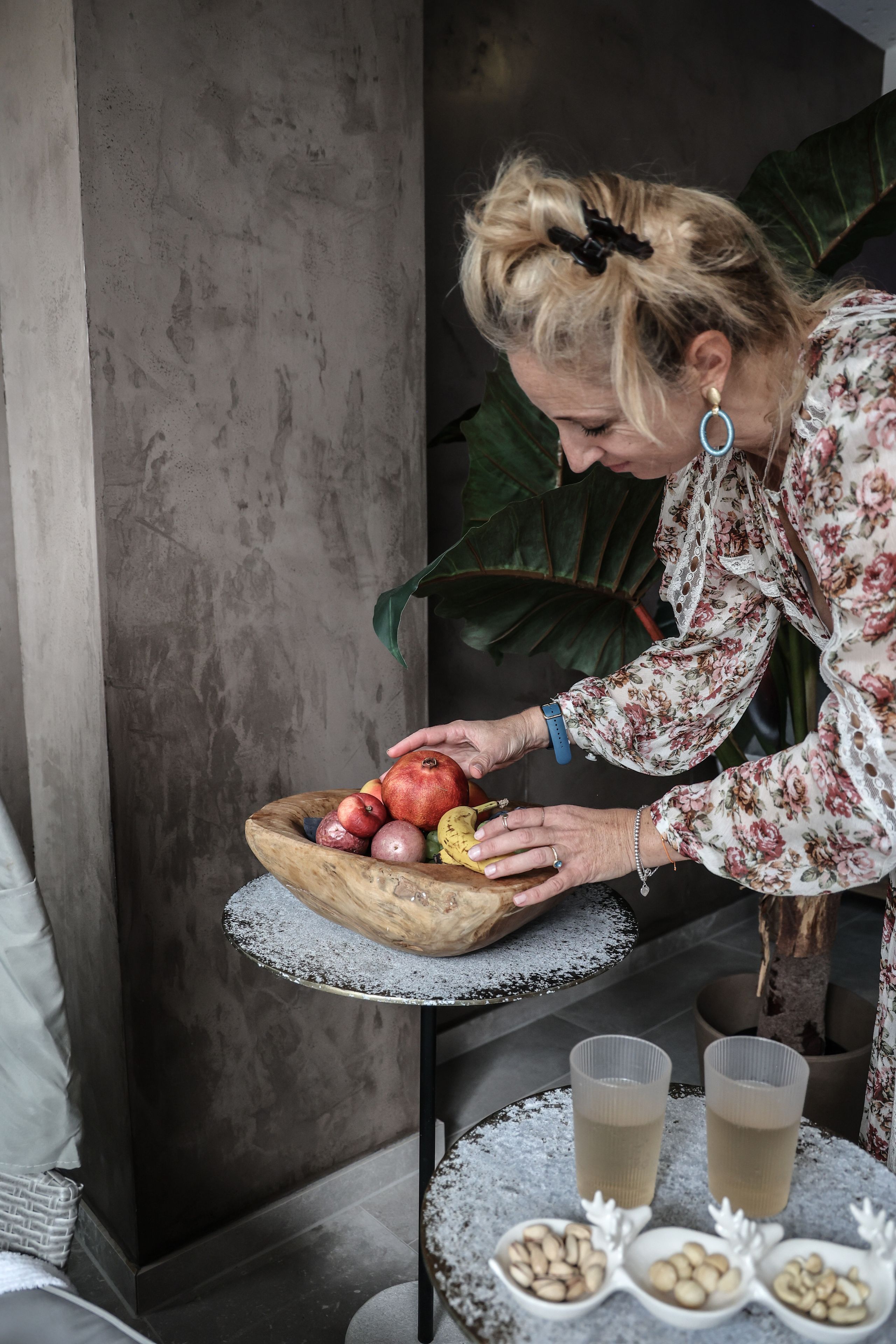 A person arranging a wooden bowl of fruits on a table with a lace tablecloth.