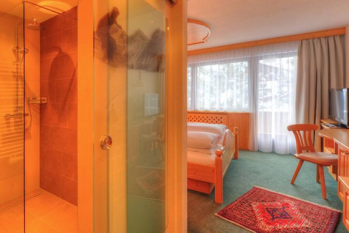 A hotel room with a glass shower enclosure, a bed with a red and white patterned rug, a wooden chair, and a television.