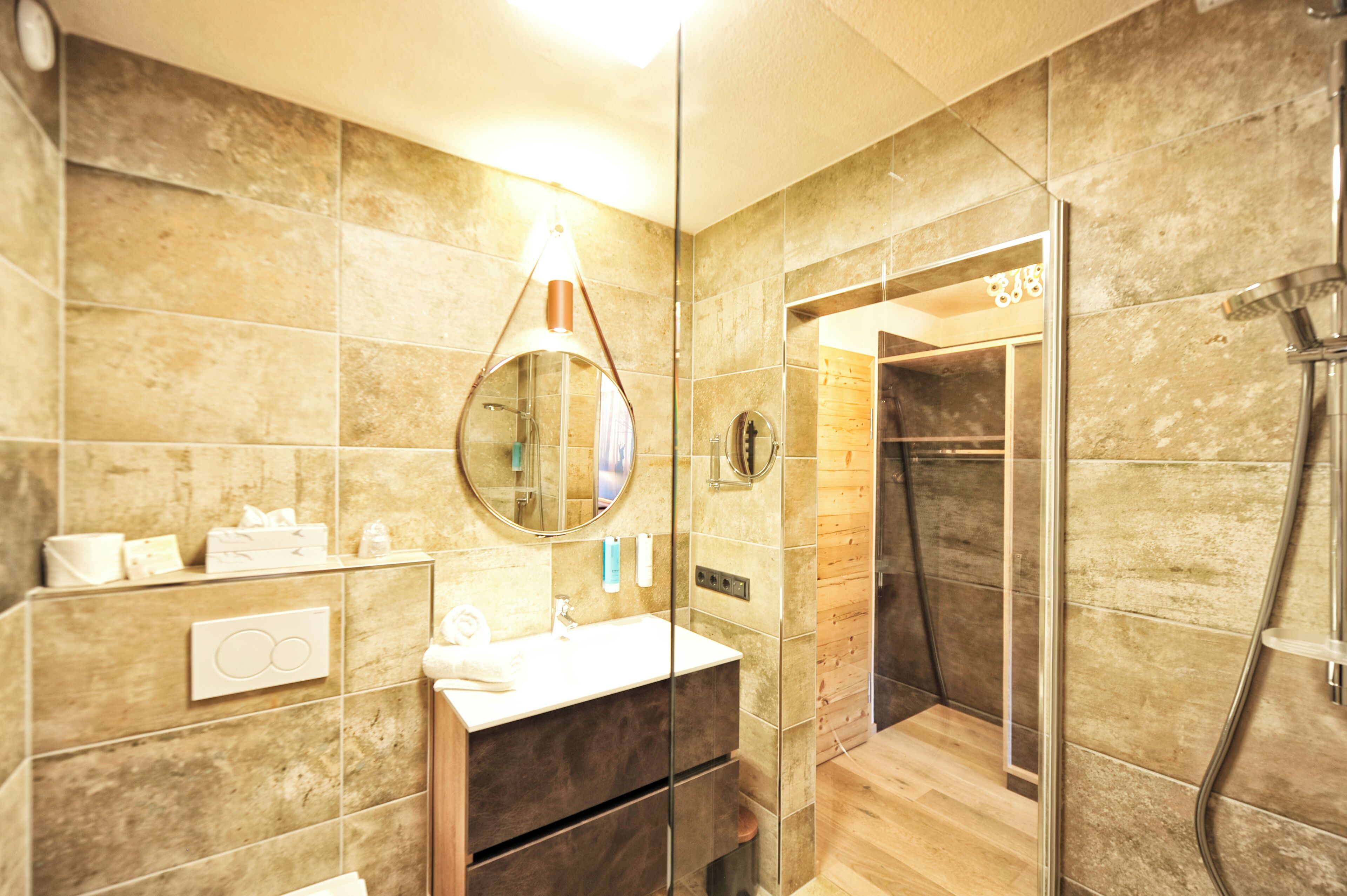 Un baño moderno con ducha envuelta en vidrio, un espejo redondo y un tocador de madera.