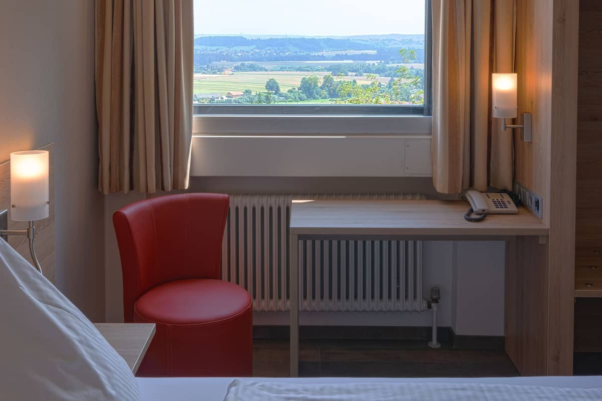 A hotel room with a bed, a desk with a telephone, a red chair, and a view of the countryside through a window.