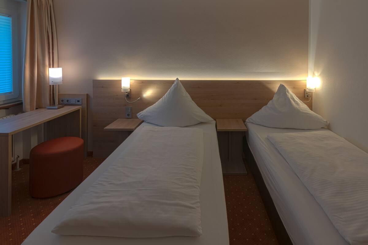 A hotel room with two single beds, a desk, a red ottoman, and bedside lamps.
