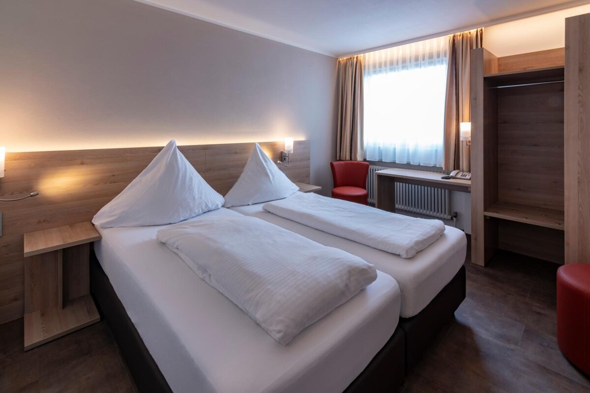 A hotel room with two beds, a red chair, a desk with a lamp, and a window with curtains.