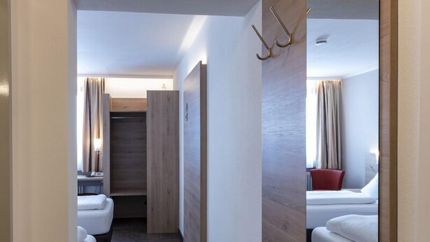 Un pasillo estrecho con un espejo grande a la derecha, que conduce a un dormitorio con cama y una silla roja.