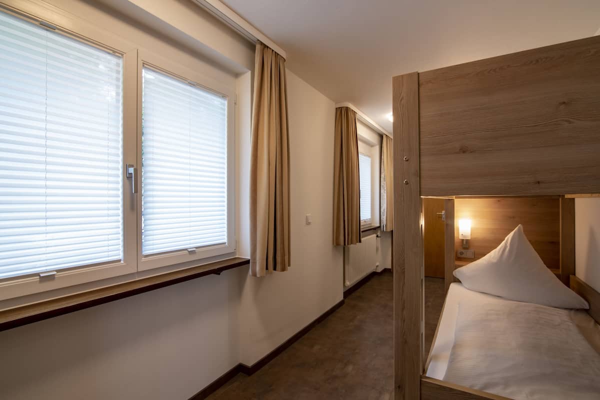 A hotel room with a bunk bed, large windows with blinds, a wooden headboard, and a nightstand with a lamp.