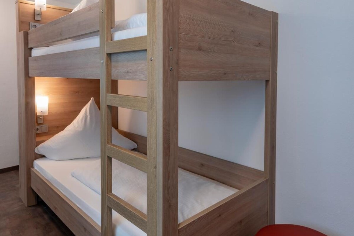 A bunk bed with a wooden frame and white bedding, a red ottoman, and a wall-mounted light fixture.