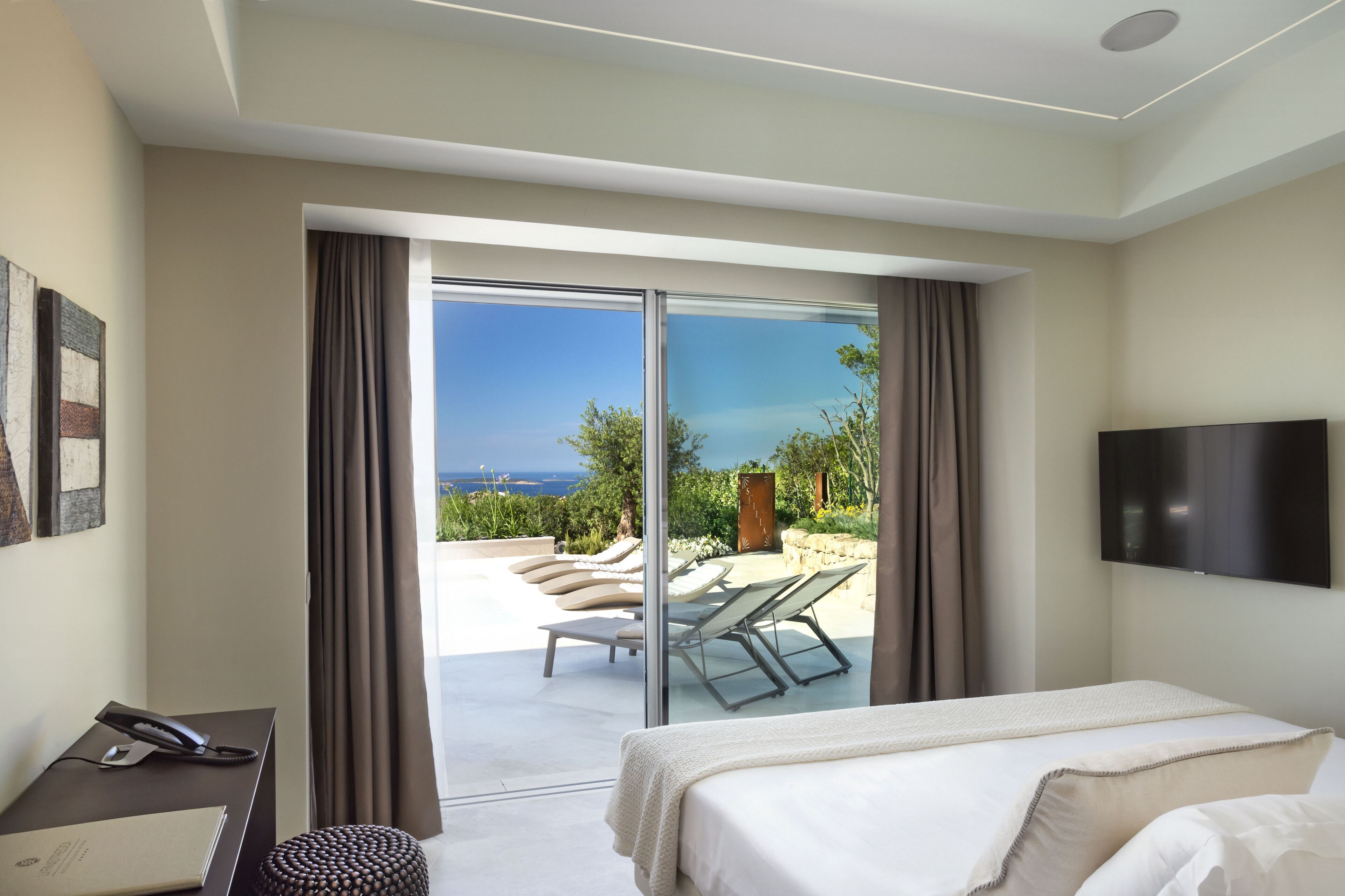 Una habitación de hotel con cama, escritorio con teléfono y televisor. Cuenta con una puerta corrediza de vidrio que da a un balcón con sillones y vistas al mar.