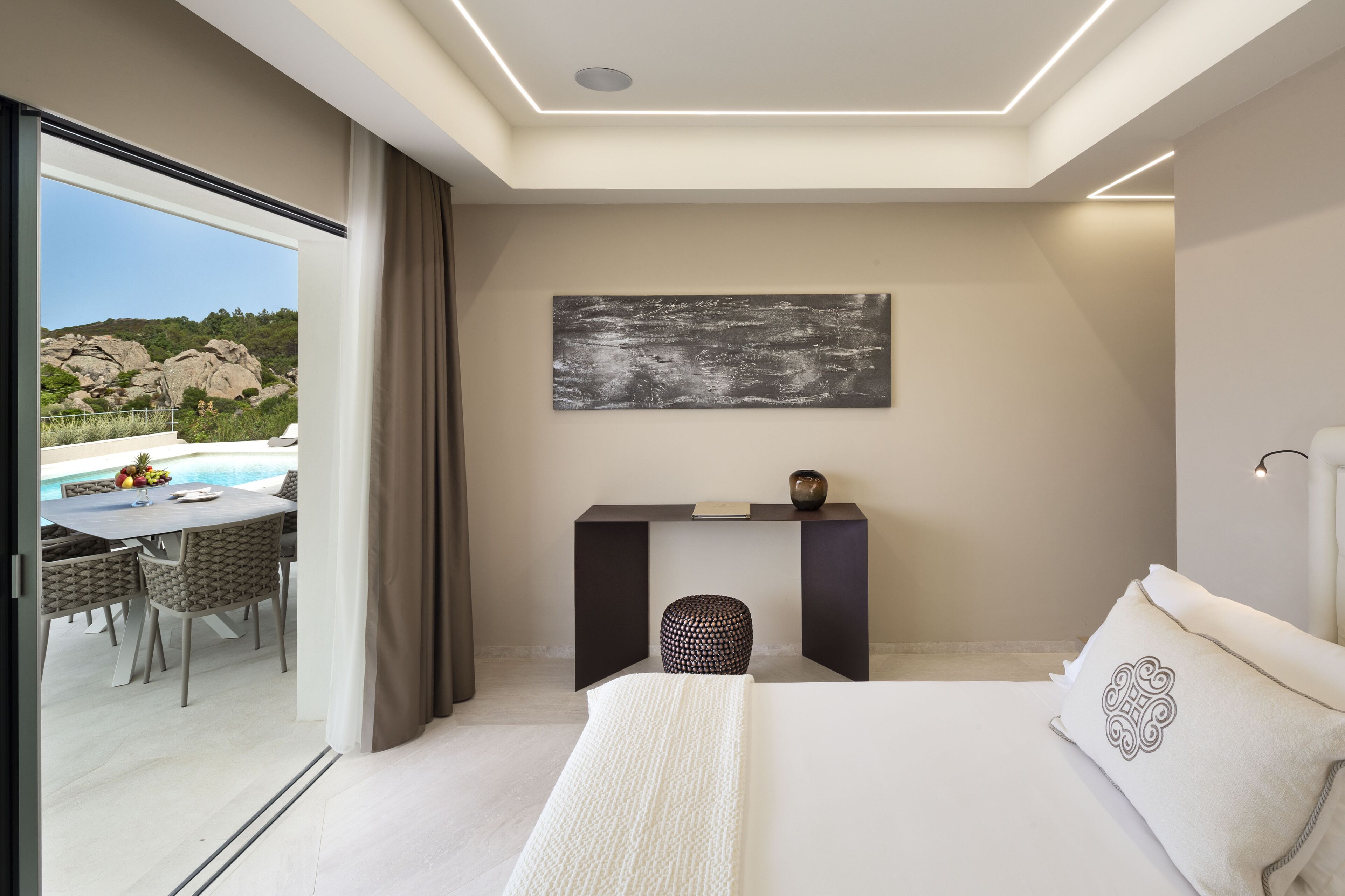 A modern bedroom with a large bed, a bedside table with a book and a decorative item, a view of a swimming pool and outdoor seating area.