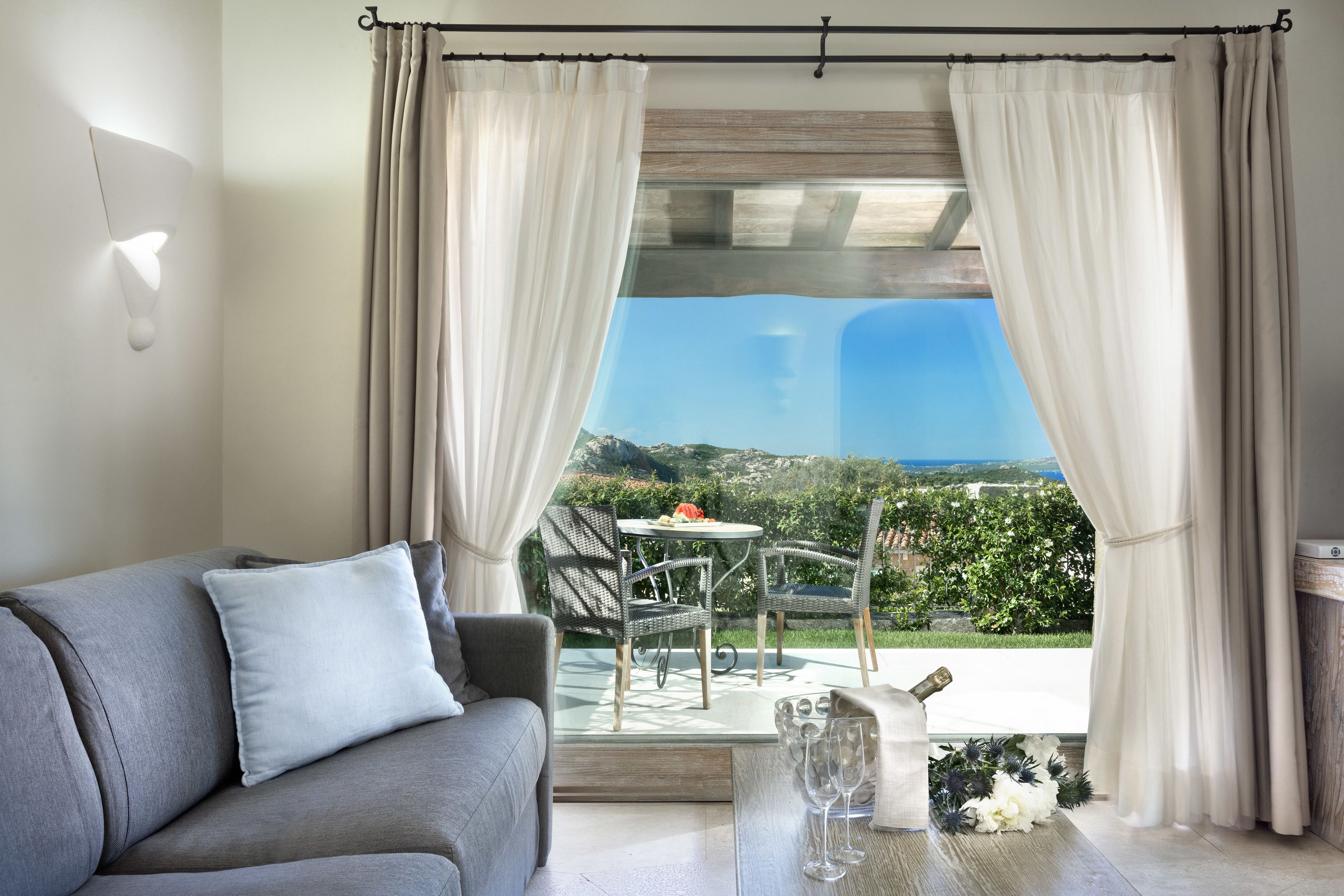A living room with a grey sofa, a table with a bottle and glasses, and a view of a patio with outdoor furniture and greenery.