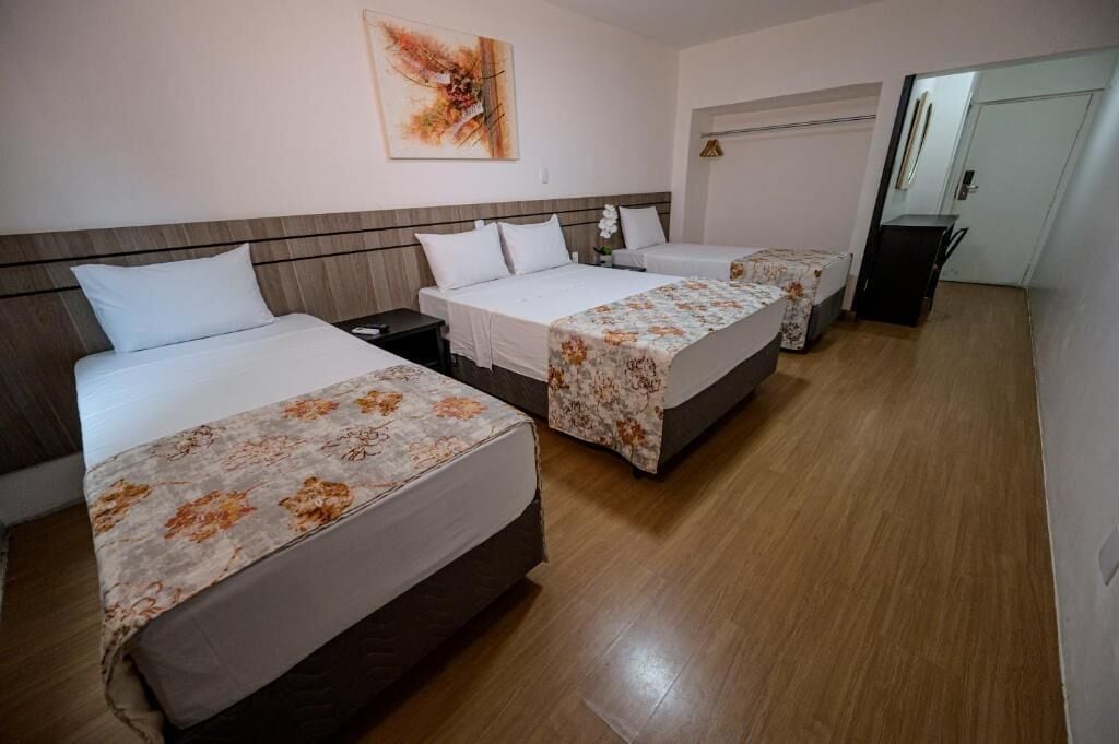 A hotel room with three single beds, each with a floral bedspread and white pillows. There is a wooden headboard, a nightstand with a lamp, and a painting on the wall.