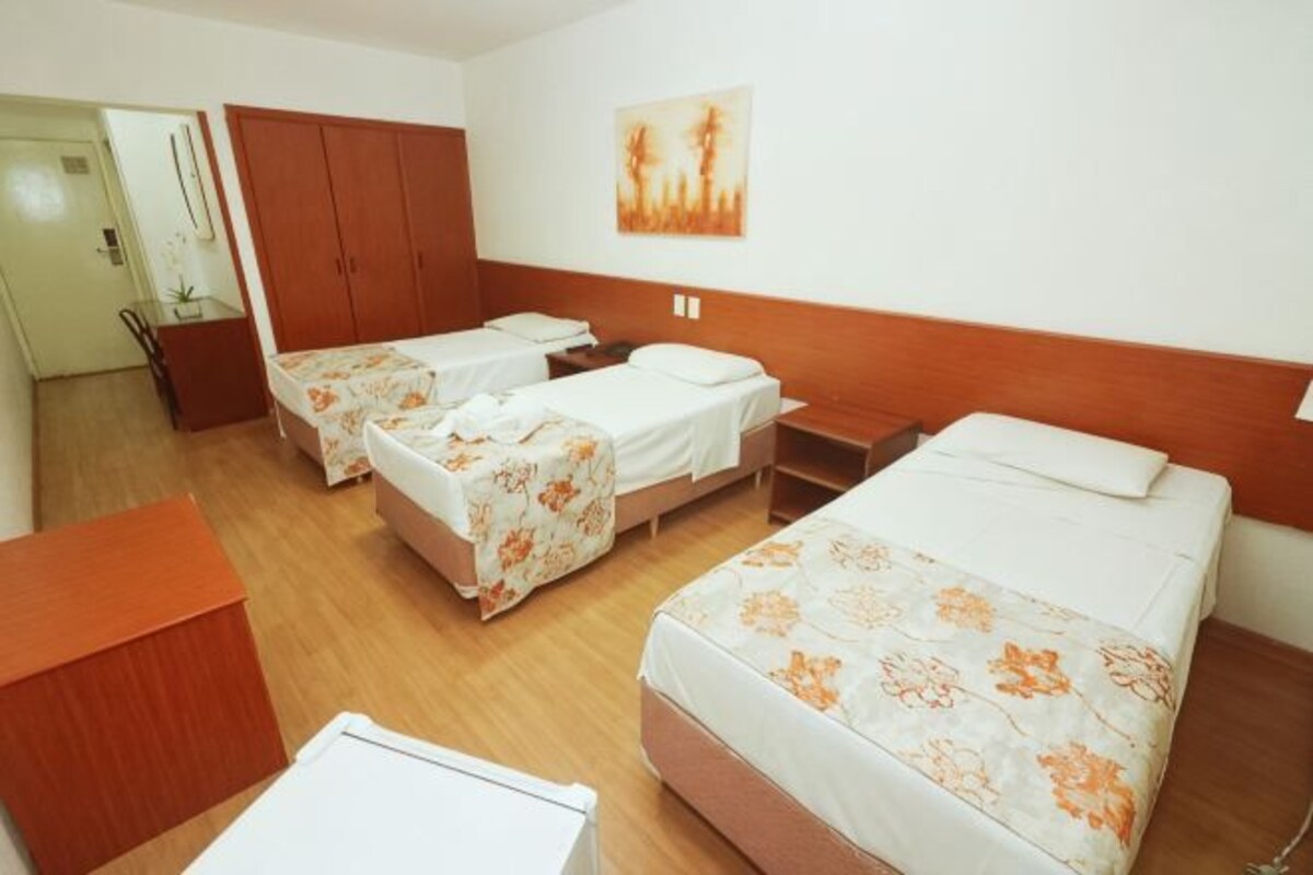 A hotel room with three beds, a wooden wardrobe, a desk, and a painting on the wall.