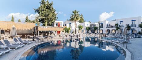 A pool area with lounge chairs and umbrellas, surrounded by white buildings with blue accents and palm trees.