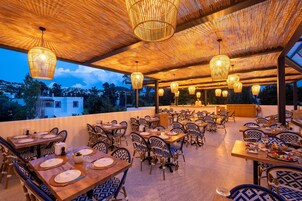 Un espacio exterior para comer con varias mesas y sillas, candelabros de mimbre colgantes y vistas al paisaje.