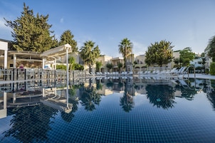 Piscina con sillones de descanso y un área de estar cubierta, rodeada de palmeras y edificios.