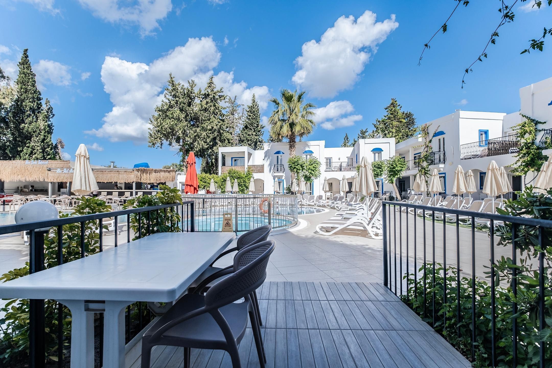 A poolside area with a table and chairs, a pool, and white buildings with blue accents in the background.