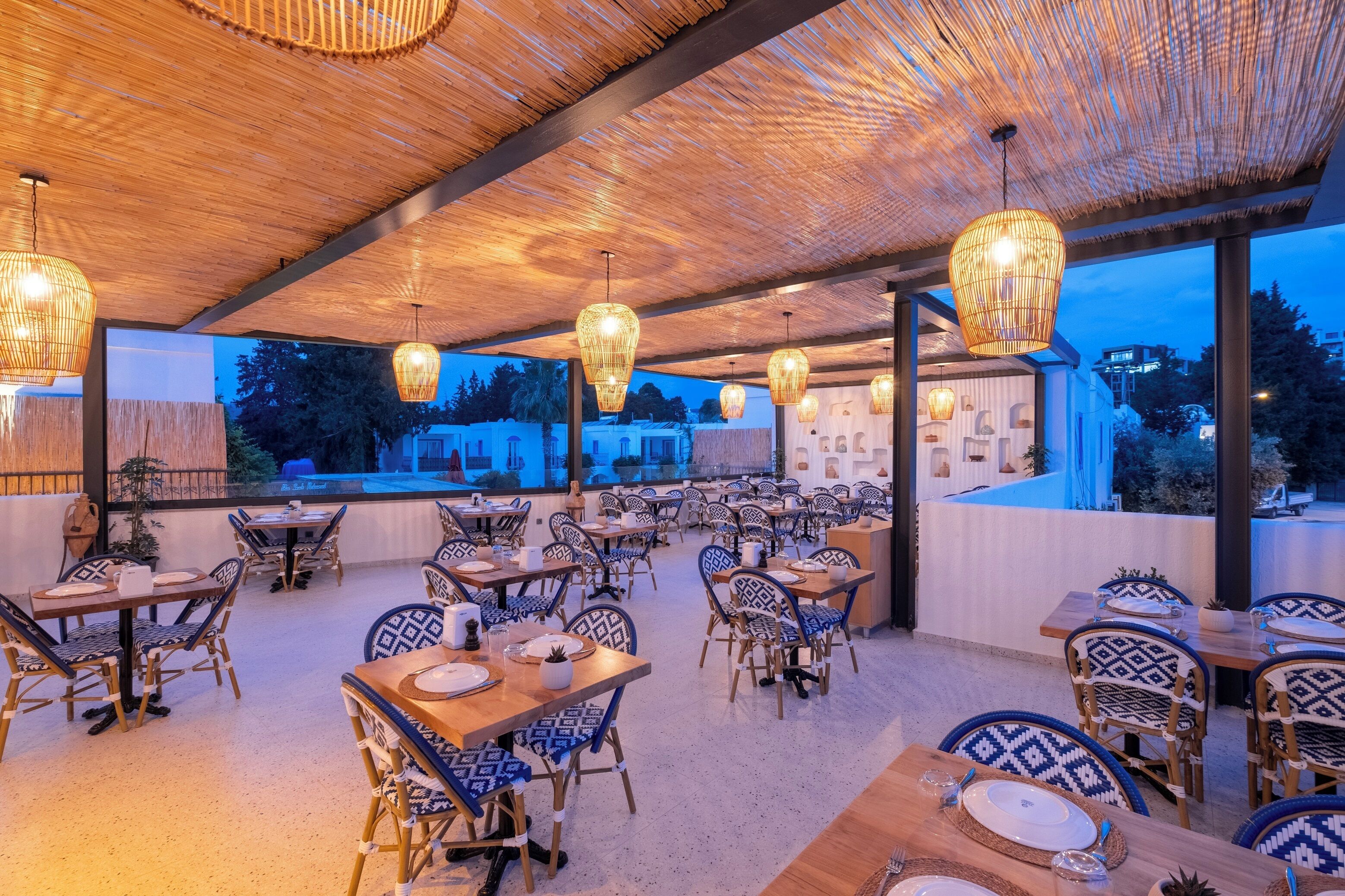 An outdoor dining area with bamboo ceiling and hanging lights, tables with blue and white patterned chairs, and a view of the cityscape at dusk.