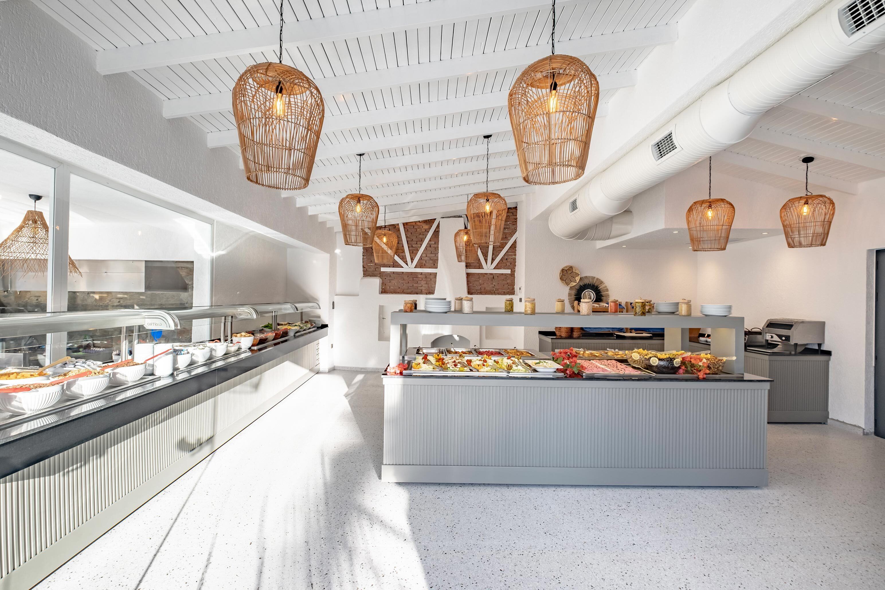 Un área de bufé moderna con iluminación colgante de mimbre, una estación central de servicio y una cocina al fondo.