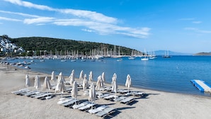 A beach with rows of sun loungers and umbrellas, a marina with boats, and a hillside in the background. - Paloma Family Club  (Bodrum)
