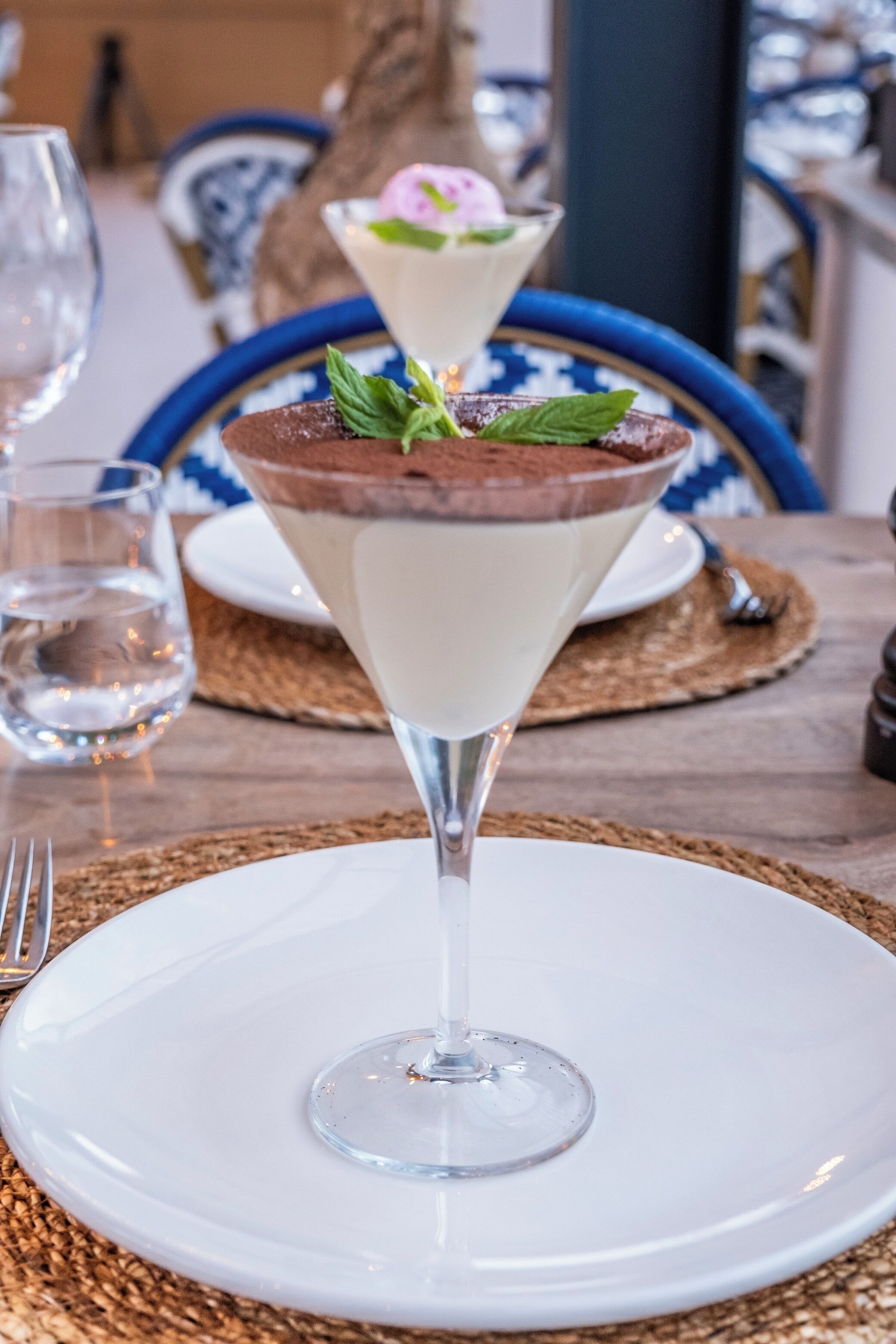 A dessert in a martini glass on a white plate, with a blue and white patterned tablecloth and a woven placemat underneath.