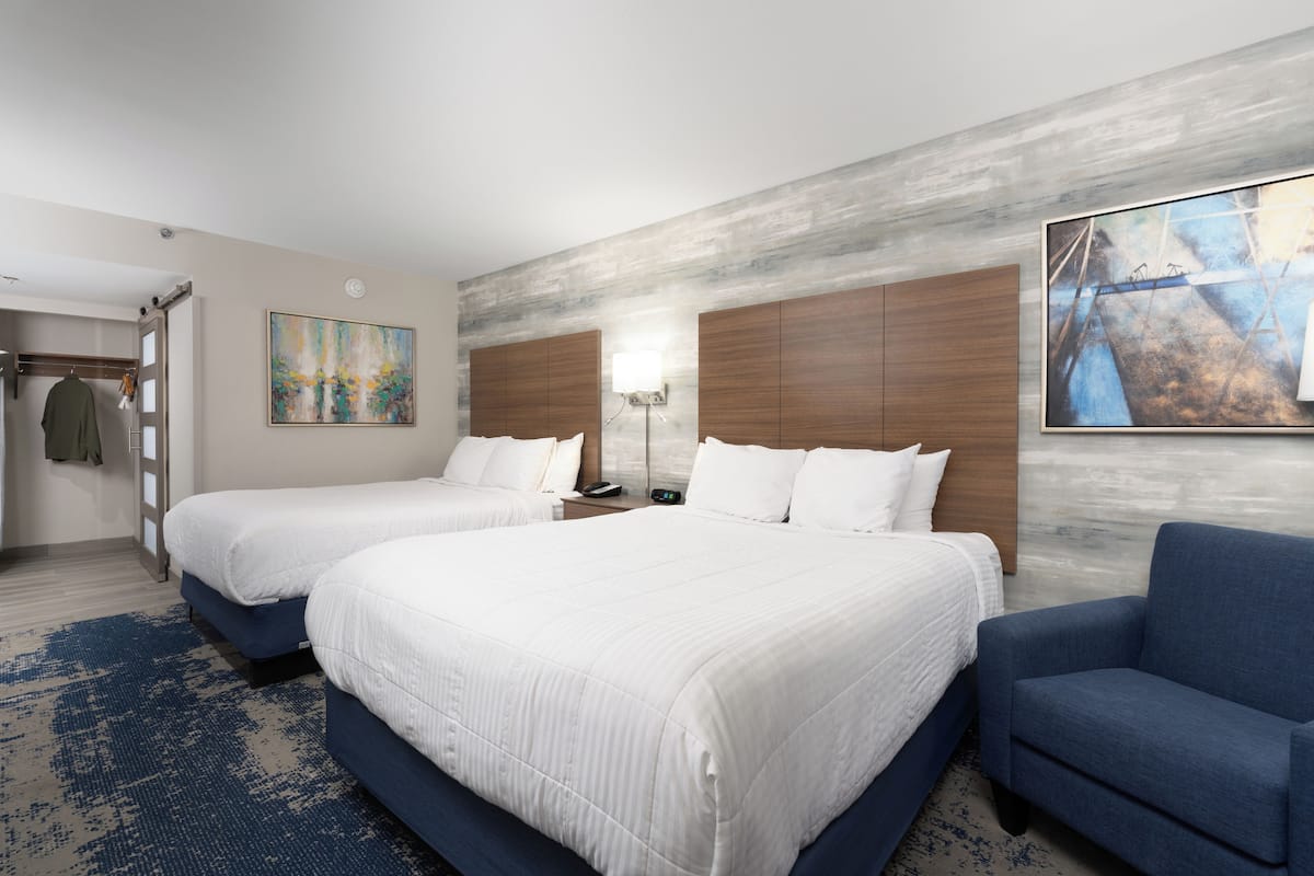 Habitación de hotel con dos camas, un sillón azul, un cuadro en la pared y un cabecero de madera.
