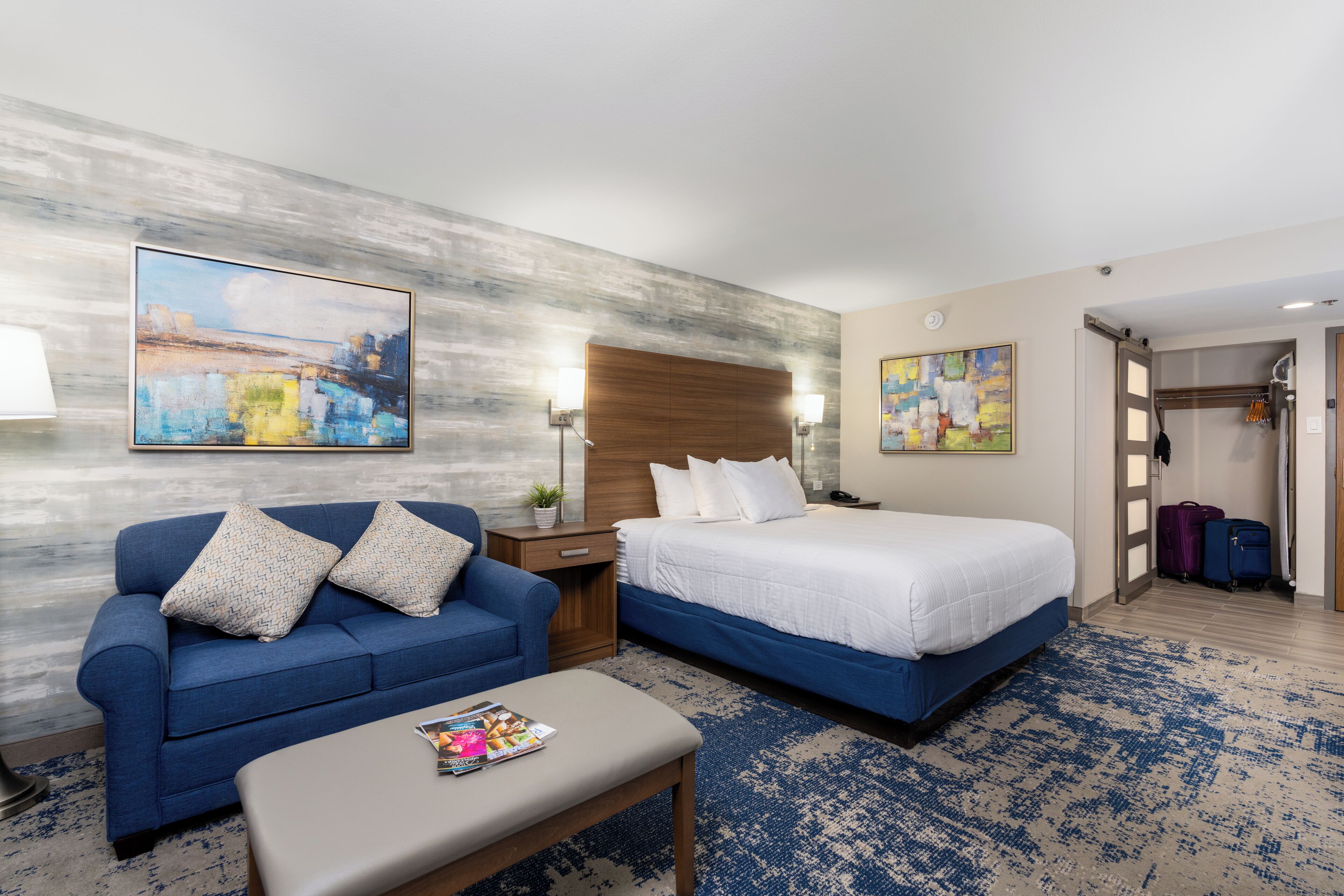 A hotel room with a bed, a sofa, a nightstand, and a table. There are two paintings on the walls and a suitcase in the corner.