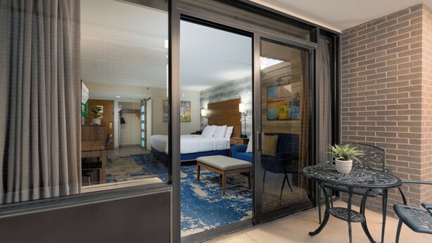 A hotel room with a large sliding glass door leading to a balcony, a bed with a blue and white patterned rug, a small table with a potted plant, and a brick wall. - MCM Elegante Hotel and Conference Center Odessa (Odessa)