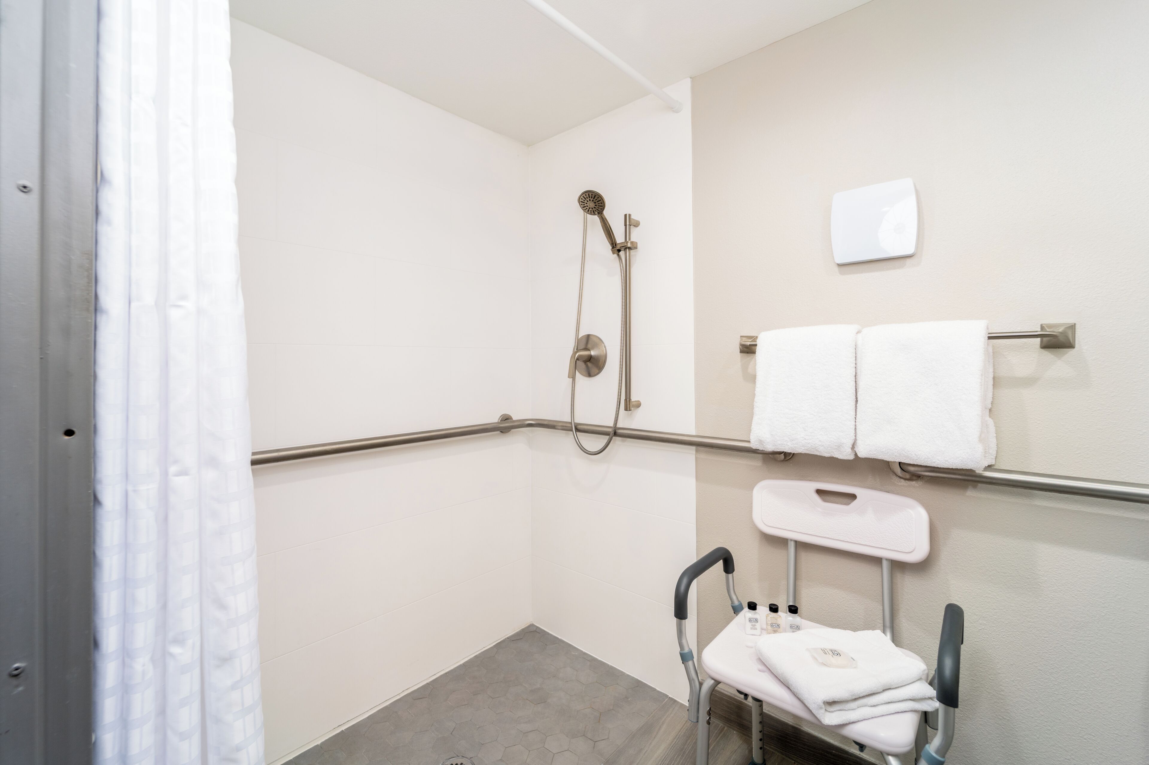 A bathroom with a shower, grab bars, a wheelchair-accessible bench, and a towel rack.