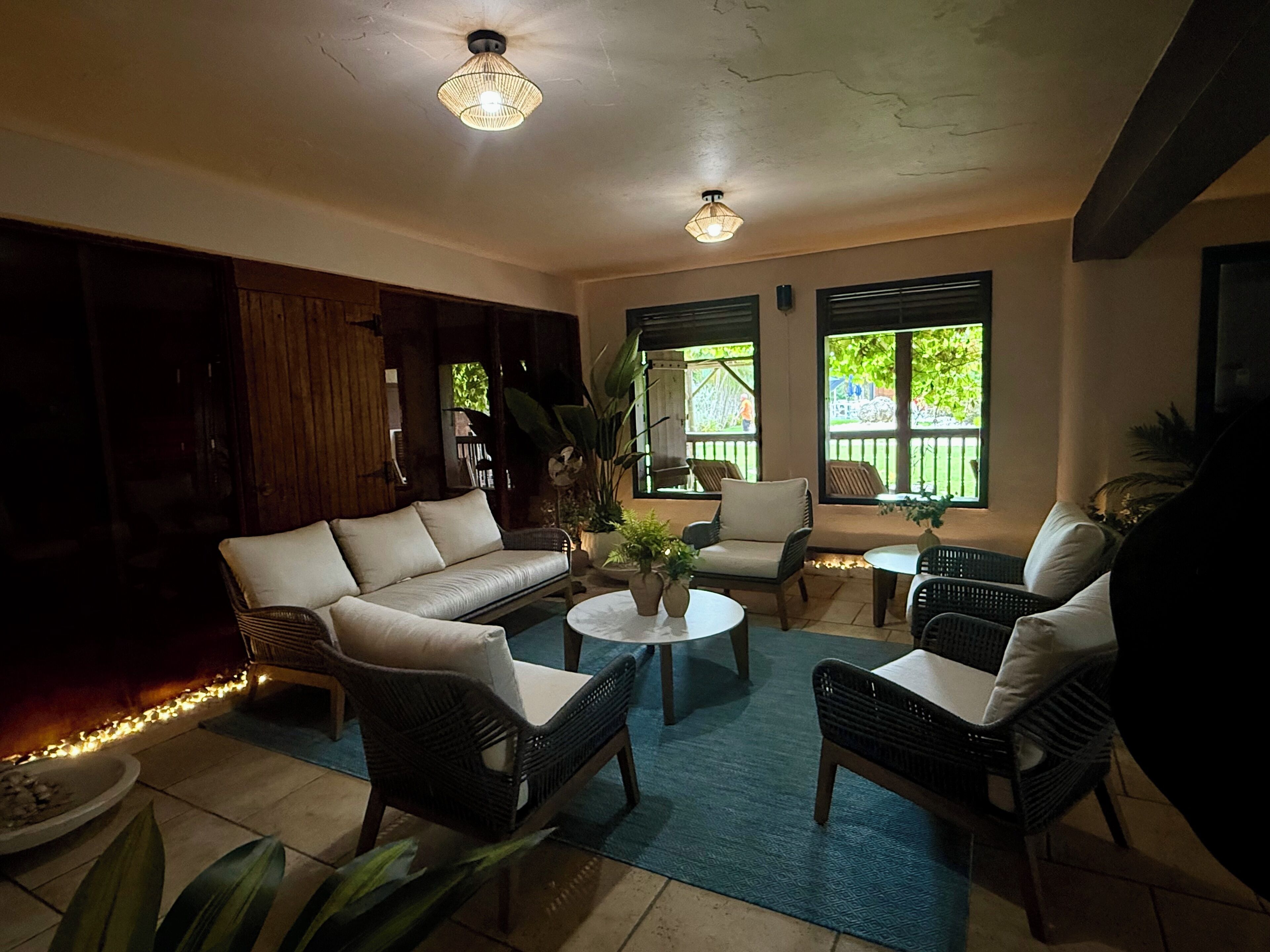 A cozy indoor seating area with a sofa, two armchairs, and a coffee table. There are potted plants and a view of the outdoors through large windows.