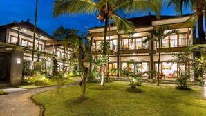 Um hotel com vários andares e varandas, rodeado por jardins bem cuidados e palmeiras.