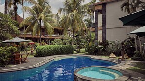 Piscina com hidromassagem, rodeada por palmeiras e um edifício com telhado de azulejos.