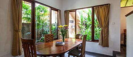 A dining area with a wooden table and chairs, a vase with flowers, and large windows with curtains.